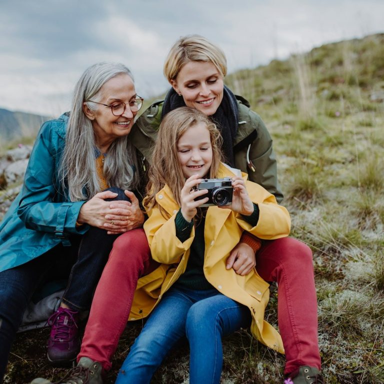 Drei Generationen sitzen gemeinsam in der Natur und betrachten eine Kamera.